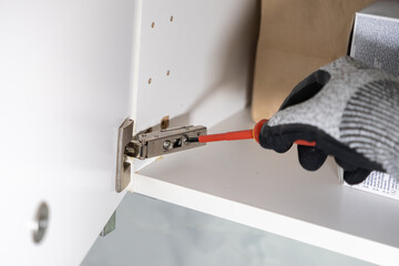 Person's gloved hand uses a red screwdriver to expertly install or adjust a metal hinge on a white kitchen cabinet, symbolizing practical home renovation and maintenance