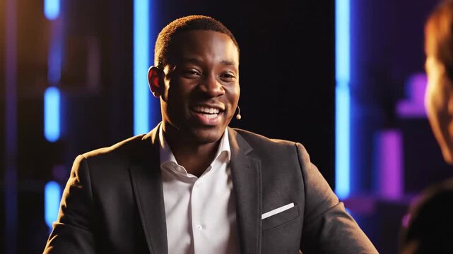 A Black man in a suit smiling during a conversation, with a blurred background featuring blue and purple lights