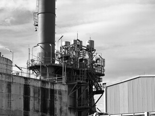 industrial landscape featuring a massive smokestack and complex piping systems at a factory. The high-contrast black and white photography highlights the weathered concrete walls, metallic scaffolding