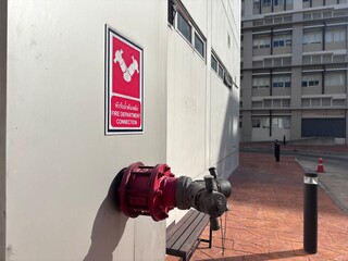 A professional close-up of a red Siamese fire department connection valve on a building wall. This shot includes clear safety signage in English and Thai, emphasizing fire protection systems, emergenc