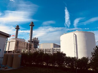 A wide-angle shot of a modern industrial power plant under a vibrant blue sky. The facility features tall smokestacks, a large white storage tank, and complex infrastructure. This high-quality image r
