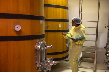 Black female wine making worker working and checking quality from wine fermenting in oak wood barrel underground room