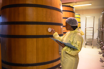 Black female wine making worker working and checking quality from wine fermenting in oak wood barrel underground room
