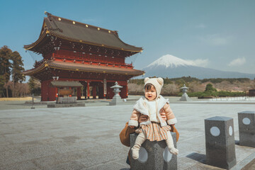 Portrait of a happy toddler girl wearing a winter bear ear hat sitting on a stone pillar with a traditional red Japanese temple gate and Mount Fuji in the background