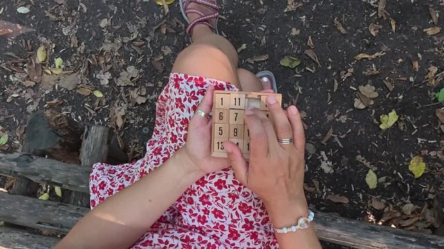 Close-up of woman playing a wooden sliding puzzle while sitting on a bench in the forest. Mindful leisure activity outdoors, slow living, relaxation, focus and calm connection with nature