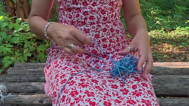 Close-up of woman untangling blue string while sitting on a wooden bench in the forest. Mindful hand activity outdoors, patience and focus, slow living lifestyle and calm connection with nature