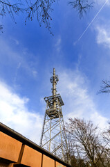 A solitary transmission tower stands tall against a vast expanse of blue sky, dotted with thin clouds and a distant airplane trail