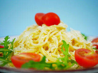 Closeup side view of spaghetti topped with grated parmesan, cherry tomatoes and arugula on white plate against blue background, appetizing Italian pasta salad detail. Pro Photo
