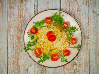 Top view of spaghetti with grated parmesan, arugula leaves and cherry tomato halves arranged in circle on white plate over rustic wooden table, Italian pasta salad