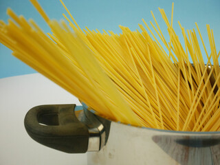 Uncooked spaghetti pasta leaning out of a metal pot with handle on blue and white background, simple Italian food ingredient image for cooking themes and kitchen design