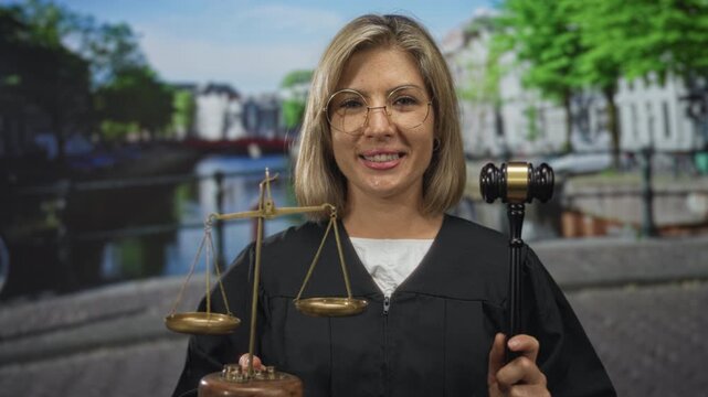Young blonde woman in black robe holding scales on street near canal water smiling; justice fairness seriousness.