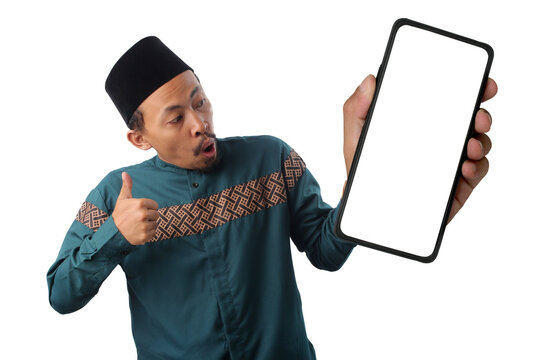 Excited Indonesian Muslim man wearing a green baju koko and black peci (songkok) presenting a phone with a blank white screen while giving a thumbs-up at camera, isolated on a white background. - Powered by Adobe