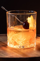 whiskey cocktail served on the rocks garnished with an orange peel anda cherry with dramatic lighting served on a wooden surface.