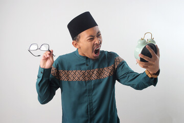 Frustrated Asian Muslim man in a green traditional outfit screaming at alarm clock in his hand, Isolated on a white background. Time stress, waking up late, or frustration with schedules and deadlines