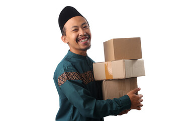 Joyful Asian Muslim man smiling while carrying a stack of cardboard boxes, symbolizing delivery, shipping, online shopping, or gift-giving, isolated on a white background