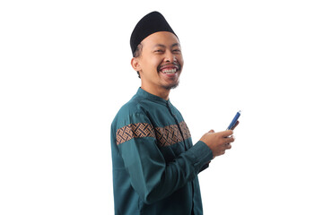 Smiling Indonesian Muslim man in a green baju koko and black peci holding his phone with a joyful expression, isolated on a white background