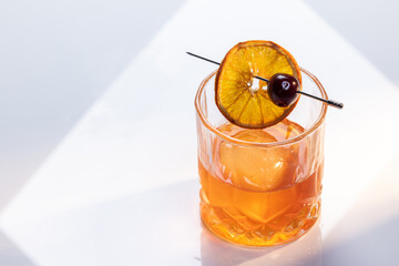 Close up of a manhattan cocktail served with one large ice cube on a reflective white surface with dual lighting tones and high contrast lights adding drama.