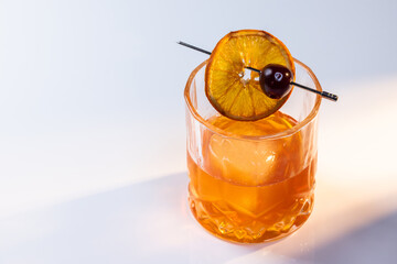 Close up of a manhattan cocktail served with one large ice cube on a reflective white surface with dual lighting tones and high contrast lights adding drama.