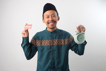 Asian Muslim man in green traditional outfit holding a credit card and alarm clock. Concept of online shopping, digital payment, and time management during Ramadan. Isolated on white background.