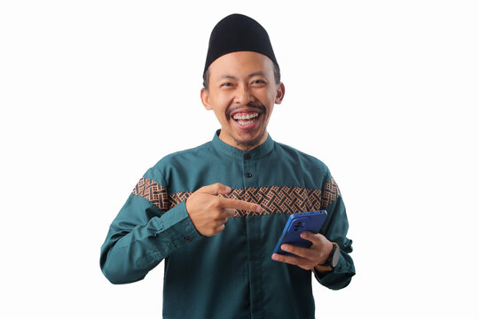 Asian Muslim man in a green traditional outfit pointing at the phone he’s holding, as if recommending something, Isolated on white background. Concept of app promotion, or online suggestion - Powered by Adobe