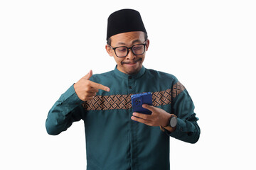 Asian Muslim man wearing a green traditional outfit and eyeglasses is pointing while holding a phone, Isolated on a white background