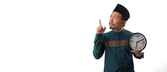 Pensive Asian man dressed in a green traditional Muslim outfit, holding a clock while looking to the side at copy space. Thinking of questions during Ramadan fasting, Isolated on a white background