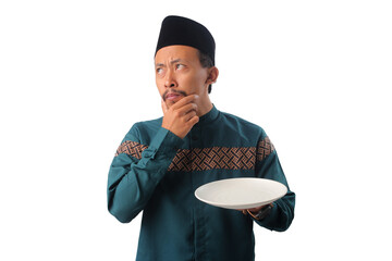 Serious Asian Muslim man wearing traditional Indonesian Muslim clothing and a black peci cap (songkok), holding an empty white plate with a thoughtful expression, isolated on a white background