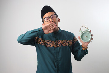 Sleepy Asian Muslim man in a green traditional outfit yawning while holding alarm clock in his hand, Isolated on a white background. Concept of tiredness, suhoor time, or lack of sleep during Ramadan