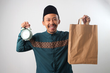 Asian Muslim man smiling at the camera while holding a brown paper bag and an alarm clock, Isolated on a white background. Food delivery, Ramadan preparation, or suhoor or iftar meal time concept