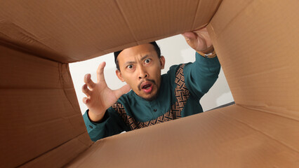 Asian Muslim man looking surprised while unpacking and opening a carton box, peeking inside with excitement, representing package delivery, online shopping, or surprise, isolated on a white background