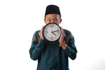 Asian Muslim man holding a wall clock in front of his face, hiding behind it with a surprised expression, Isolated on a white background, Concept of time pressure, delay, or missing a deadline.