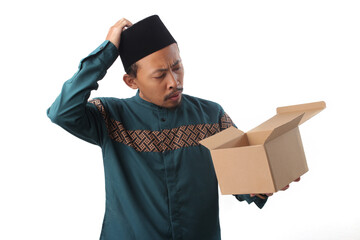 Unhappy Asian Muslim man unboxing his new package from online shopping, looking inside the box with a surprised and disappointed expression, isolated on a white background