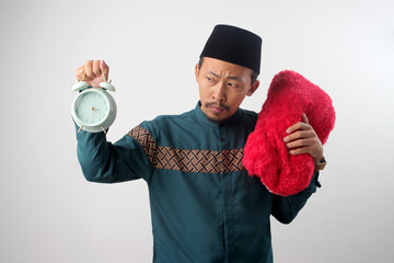 Displeased Asian Muslim man in a green traditional outfit holding a red pillow and an alarm clock, looking upset. Isolated on a white background.