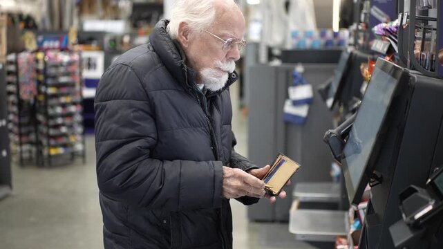 An elderly man stands at a self checkout in a large hardware store while shopping for tools and supplies during a busy afternoon.