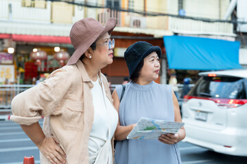 Two senior asian female travelers standing on city sidewalk holding map discussing direction during retirement journey exploring cultural destination lifestyle vacation friendship outdoor travel