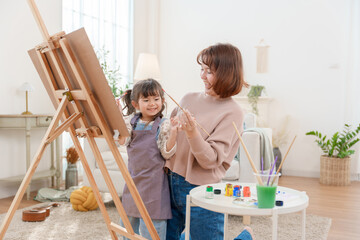 Cheerful Asian mother and her little daughter laughing while painting on an easel at home, enjoying creative art hobby and family togetherness