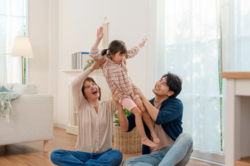 Asian parents sitting on floor, lifting excited daughter high in bright living room. Family sharing playful, heartwarming moment of love and fun together.