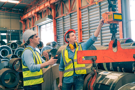 Caucasian industrial engineer men controlling overhead crane with remote in factory. Worker partner wear safety helmet vest lifting heavy metal coil material. Manufacturing hoist machine logistics