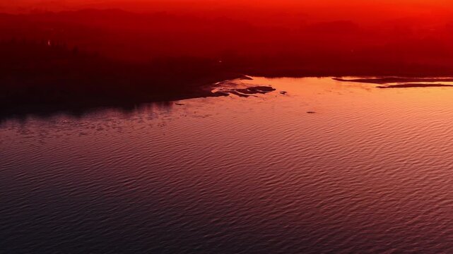 Red dusk reflections on lake surface. Close view of rippled water tinted crimson, dark treeline in distance.