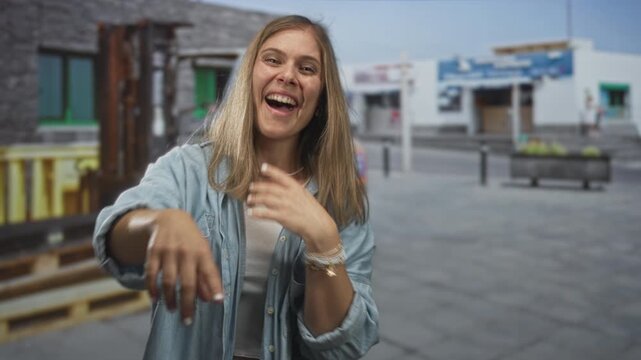 Young blonde woman points finger to camera while covering mouth in laughter on urban street; amusement.
