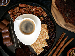 A cup of black coffee on a saucer, surrounded by coffee beans, nuts, chocolate, and sesame cookies.