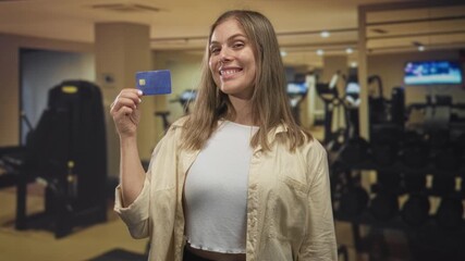 Young blonde woman showing and pointing finger to creditcard in modern well lit gym studio; confidence.