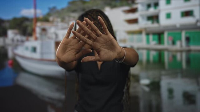 Woman raising hands to block face at port pier, palms facing camera; shyness privacy avoidance retreat.