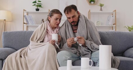 Sick couple at home with fever and tissues. Wrapped in a blanket, they sip tea and consider medicine on the sofa, battling virus illness symptoms and chills. Theme: home flu care and recovery.