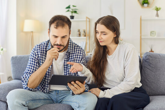 Couple with tablet browsing internet at home. Sitting on sofa, they search together, compare options, and plan purchases in app in cozy living room. Online shopping and technology. 