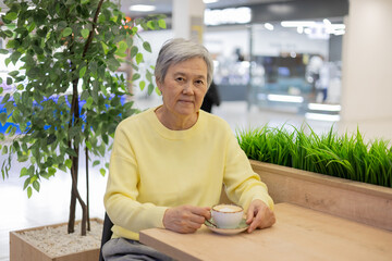 Elderly Asian woman with a cup of coffee in a cafe in a shopping mall, happy pensioner in a cafe