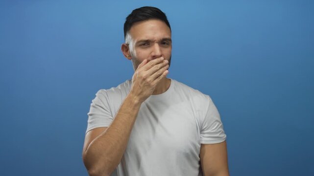 Young man covers mouth in surprise against a blue background, expressing shock and disbelief.