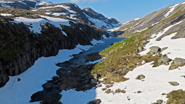 Aerial view of alpine river along the Kjerag hiking trail Norway. Drone shot flying above a narrow stream between snow patches and rocky slopes on the way to Kjerag during a clear summer day.