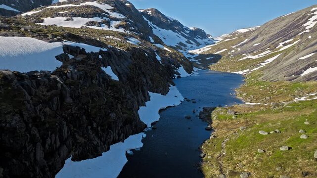 Aerial view of alpine river and rocky valley near Kjerag. Drone footage showing flowing water, snow patches, and steep mountain sides in remote hiking terrain.
