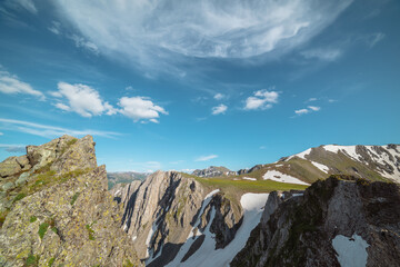 Aerial dizzying view to rocky cliff between sharp rocks under clouds in blue sky. Vertigo scenery with sheer crags above precipice, snowy ridge and mountain top far away in cloudy changeable weather. © Daniil